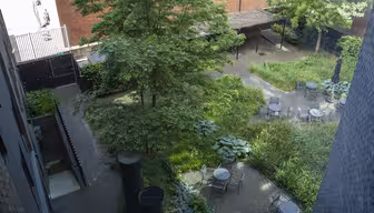 Udsigt over en anlagt gårdhave med flere borde med stole, omgivet af frodige grønne planter og træer. Området er brolagt med mursten og omkranset af murstensbygninger, hvilket skaber en fredfyldt udendørs siddeplads.