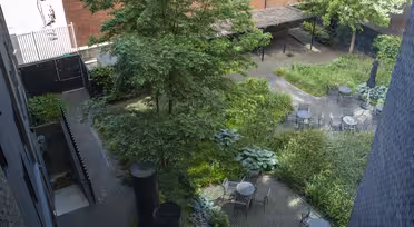 Udsigt over en anlagt gårdhave med flere borde med stole, omgivet af frodige grønne planter og træer. Området er brolagt med mursten og omkranset af murstensbygninger, hvilket skaber en fredfyldt udendørs siddeplads.