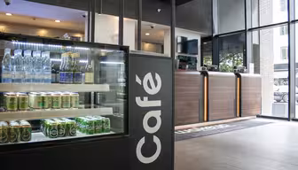 En moderne café med et glasdisplay til venstre med flaskedrinks og snacks. Skranken til højre har en lækker træfinish. Et stort vindue i baggrunden lukker naturligt lys ind og afslører udendørs grønt.