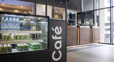 En moderne café med et glasdisplay til venstre med flaskedrinks og snacks. Skranken til højre har en lækker træfinish. Et stort vindue i baggrunden lukker naturligt lys ind og afslører udendørs grønt.