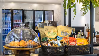 En udstilling af snacks på en café omfatter croissanter under en glaskuppel, poser med chips og kurve med slik og jordnødder. Kølede drikkevarer og flasker på hylder er i baggrunden med hængende planter over.