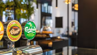 Nærbillede af ølhaner med Tuborg Classic- og Carlsberg-logoer i moderne bar-miljø. Baggrunden viser sløret grønt og moderne indretning med hængende lys.