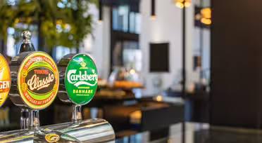 Nærbillede af ølhaner med Tuborg Classic- og Carlsberg-logoer i moderne bar-miljø. Baggrunden viser sløret grønt og moderne indretning med hængende lys.