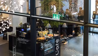 Se gennem glas af en moderne cafe med folk, der bestiller ved skranken. Cafeen har kager, flasker og et "Café"-skilt. Indretningen inkluderer hængende lys og planter. Et nødudgangsskilt er synligt ovenfor.