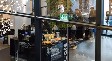 Se gennem glas af en moderne cafe med folk, der bestiller ved skranken. Cafeen har kager, flasker og et "Café"-skilt. Indretningen inkluderer hængende lys og planter. Et nødudgangsskilt er synligt ovenfor.
