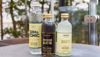 Tre cocktails på flaske på et bord: Gin & Tonic, Espresso Martini og Mojito. Hver har karakteristiske etiketter i bløde farver med tekstdetaljer, der beskriver smag. Flaskerne er sat mod en sløret udendørs baggrund af grønt.