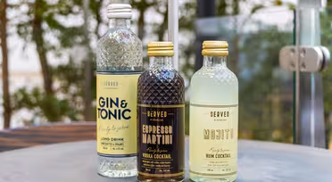 Tre cocktails på flaske på et bord: Gin & Tonic, Espresso Martini og Mojito. Hver har karakteristiske etiketter i bløde farver med tekstdetaljer, der beskriver smag. Flaskerne er sat mod en sløret udendørs baggrund af grønt.