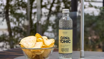 En flaske gin og tonic står på et bord ved siden af en glasskål fyldt med kartoffelchips. Indstillingen er udendørs med sløret grønt i baggrunden.