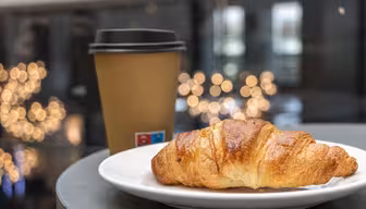 En flaget croissant sidder på en hvid tallerken ved siden af en brun engangs kaffekop med sort låg. Den bløde bokeh-baggrund har varme gyldne lys, hvilket skaber en hyggelig atmosfære.