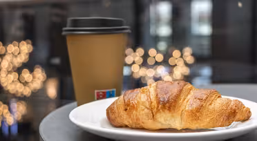 En flaget croissant sidder på en hvid tallerken ved siden af en brun engangs kaffekop med sort låg. Den bløde bokeh-baggrund har varme gyldne lys, hvilket skaber en hyggelig atmosfære.