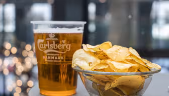 En plastikkop Carlsberg-øl står ved siden af en glasskål fyldt med kartoffelchips på et bord. Baggrunden er sløret med ude af fokus lys, der skaber en hyggelig atmosfære.