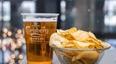 En plastikkop Carlsberg-øl står ved siden af en glasskål fyldt med kartoffelchips på et bord. Baggrunden er sløret med ude af fokus lys, der skaber en hyggelig atmosfære.