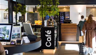 Moderne caféinteriør med digitale kiosker, en skranke med et kasseapparat og et køleskab fyldt med drikkevarer. Grønne planter hænger fra loftet. Et par kunder står ved disken, og stemningen er lys og indbydende.