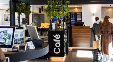 Moderne caféinteriør med digitale kiosker, en skranke med et kasseapparat og et køleskab fyldt med drikkevarer. Grønne planter hænger fra loftet. Et par kunder står ved disken, og stemningen er lys og indbydende.