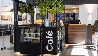 Moderne café med en disk med kager, snacks og kaffeposer. På den sorte skiltning står der "Café". Fra loftet hænger grønne planter, og caféen ligger ved siden af en glasindgang med synlige siddepladser indenfor.