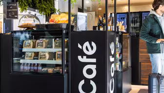 Et moderne caféinteriør med en udstilling af pakkede snacks, vinflasker og desserter under en glaskuppel. En kunde med en kuffert er til højre, delvist synlig. Caféen har sorte diske og grønt over hovedet.