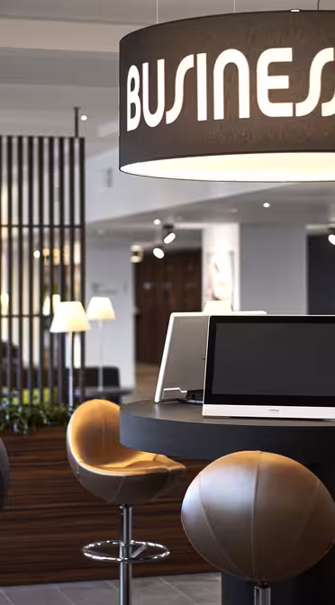 Moderne businesscenter med slanke sorte borde og runde stole. Computere vises på borde under store hængende lamper med teksten "Business Center". Træaccenter og grønt skaber en professionel atmosfære.