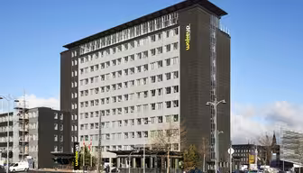 En moderne hotelbygning i flere etager med en grå facade og store vinduer. "Wakeup" står der med gult på siden. Forgrunden viser en gade med biler, cykler og spredte fodgængere under en delvist overskyet himmel.