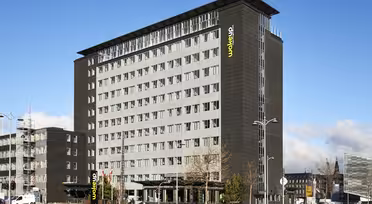 En moderne hotelbygning i flere etager med en grå facade og store vinduer. "Wakeup" står der med gult på siden. Forgrunden viser en gade med biler, cykler og spredte fodgængere under en delvist overskyet himmel.
