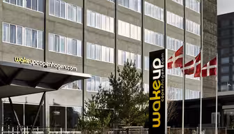 Yderside af Wakeup Copenhagen hotel med en moderne grå facade. Hotellets navn er vist med gult på et sort skilt. Tre danske flag vajer nær indgangen, og små træer og buske er i forgrunden.