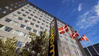 Høj, moderne bygning under en blå himmel med et mørkt lodret skilt med teksten "Wakeup Copenhagen" i forgrunden. Tre danske flag vajer på pæle i nærheden, og nogle træer er synlige i bunden af bygningen.