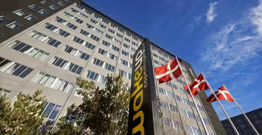 Høj, moderne bygning under en blå himmel med et mørkt lodret skilt med teksten "Wakeup Copenhagen" i forgrunden. Tre danske flag vajer på pæle i nærheden, og nogle træer er synlige i bunden af bygningen.