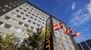 Høj, moderne bygning under en blå himmel med et mørkt lodret skilt med teksten "Wakeup Copenhagen" i forgrunden. Tre danske flag vajer på pæle i nærheden, og nogle træer er synlige i bunden af bygningen.