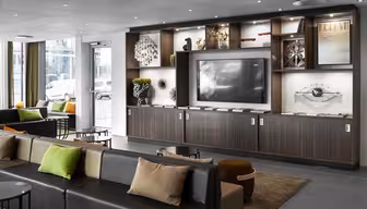Moderne hotellobby med slanke hylder i mørkt træ med dekorative genstande og en stor tv-skærm. Siddeområdet omfatter en lang sofa med farverige hynder og runde borde, alt sammen under et loft med forsænket belysning. Store vinduer har udsigt over byen.