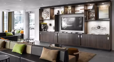 Moderne hotellobby med slanke hylder i mørkt træ med dekorative genstande og en stor tv-skærm. Siddeområdet omfatter en lang sofa med farverige hynder og runde borde, alt sammen under et loft med forsænket belysning. Store vinduer har udsigt over byen.