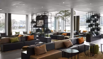 Moderne loungeområde med grå sofaer, farverige puder og runde glasborde. Naturligt lys strømmer gennem store vinduespartier. Indendørs planter på hylder tilføjer grønt, og et tæppe dækker gulvet og skaber en hyggelig atmosfære.