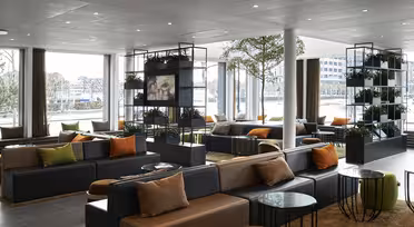 Moderne loungeområde med grå sofaer, farverige puder og runde glasborde. Naturligt lys strømmer gennem store vinduespartier. Indendørs planter på hylder tilføjer grønt, og et tæppe dækker gulvet og skaber en hyggelig atmosfære.
