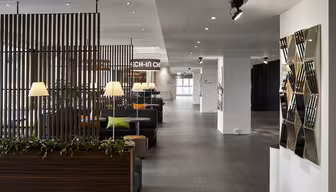 Moderne lobby med slanke træpaneldelere og planter. Bænke og borde med lamper skaber et hyggeligt opholdsområde. Geometrisk vægkunst og loftslamper tilføjer den moderne følelse. På et skilt står "CHECK-IN" i baggrunden.