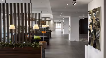 Moderne lobby med slanke træpaneldelere og planter. Bænke og borde med lamper skaber et hyggeligt opholdsområde. Geometrisk vægkunst og loftslamper tilføjer den moderne følelse. På et skilt står "CHECK-IN" i baggrunden.