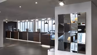 Moderne hotelobby med en elegant reception og computerstationer langs en væg. Et dekorativt spejl med geometriske sektioner afspejler det moderne design. Rummet er godt oplyst, med mørkt træ og neutrale toner.