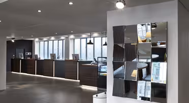 Moderne hotelobby med en elegant reception og computerstationer langs en væg. Et dekorativt spejl med geometriske sektioner afspejler det moderne design. Rummet er godt oplyst, med mørkt træ og neutrale toner.