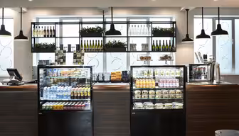 Et moderne caféinteriør med glasmontre med drinks, snacks og sandwich. Hylderne bag rummer vinflasker og planter. Pendellamper hænger fra loftet, og et kasseapparat er synligt til venstre.