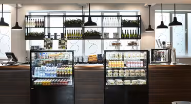 Et moderne caféinteriør med glasmontre med drinks, snacks og sandwich. Hylderne bag rummer vinflasker og planter. Pendellamper hænger fra loftet, og et kasseapparat er synligt til venstre.