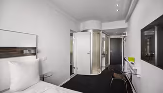 Moderne hotelværelse med et slankt design, med en seng med hvidt sengetøj, et glasskrivebord med en stol, et vægmonteret tv og en buet glasindkapslet bruser. Rummet har sort gulv og hvide vægge, hvilket skaber en minimalistisk æstetik.