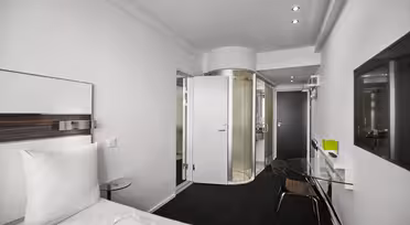 Moderne hotelværelse med et slankt design, med en seng med hvidt sengetøj, et glasskrivebord med en stol, et vægmonteret tv og en buet glasindkapslet bruser. Rummet har sort gulv og hvide vægge, hvilket skaber en minimalistisk æstetik.