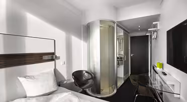 Moderne hotelværelse med en hvid seng, en gennemsigtig cylindrisk bruser, en sort stol og et glasskrivebord. Væggene er hvide, og på den ene væg er der monteret et stort fladskærms-tv. Døren og gulvet er mørke, hvilket skaber en kontrast.