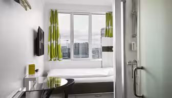 Kompakt, moderne hotelværelse med en enkeltseng under et stort vindue, med grønne og hvide gardiner. Et væghængt tv og et lille skrivebord med en stol er til venstre. Til værelset hører et bruseområde med en glasdør til højre. Bybilledet synligt udenfor.
