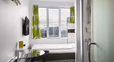 Kompakt, moderne hotelværelse med en enkeltseng under et stort vindue, med grønne og hvide gardiner. Et væghængt tv og et lille skrivebord med en stol er til venstre. Til værelset hører et bruseområde med en glasdør til højre. Bybilledet synligt udenfor.
