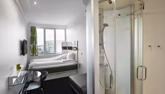 Et kompakt, moderne hotelværelse med en seng, et vægmonteret tv og en glasindkapslet bruser. Der er et lille skrivebord med en stol og en skammel. Rummet har et stort vindue med udsigt til en bygning, og et lyst grønt og hvidt gardin.