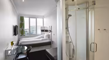 Et kompakt, moderne hotelværelse med en seng, et vægmonteret tv og en glasindkapslet bruser. Der er et lille skrivebord med en stol og en skammel. Rummet har et stort vindue med udsigt til en bygning, og et lyst grønt og hvidt gardin.