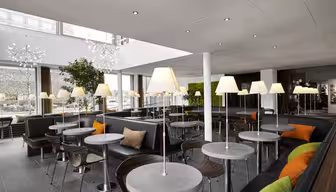 Moderne caféinteriør med slanke grå borde og stole, accentueret af livlige orange og grønne puder. Høje gulvlamper langs rummet, og store vinduer bringer naturligt lys ind. Grønt og moderne indretning forstærker den indbydende atmosfære.