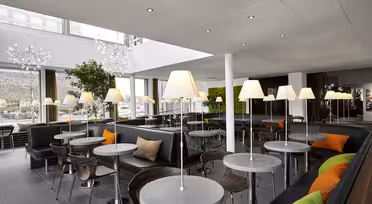Moderne caféinteriør med slanke grå borde og stole, accentueret af livlige orange og grønne puder. Høje gulvlamper langs rummet, og store vinduer bringer naturligt lys ind. Grønt og moderne indretning forstærker den indbydende atmosfære.
