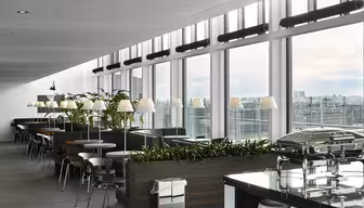 En moderne spiseplads med store vinduer med udsigt over byen. Rummet byder på sorte borde, stole, planter og flere lamper, sammen med buffetstationer og loftslamper, hvilket skaber en lys, indbydende atmosfære.