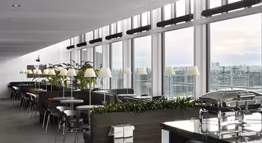 En moderne spiseplads med store vinduer med udsigt over byen. Rummet byder på sorte borde, stole, planter og flere lamper, sammen med buffetstationer og loftslamper, hvilket skaber en lys, indbydende atmosfære.