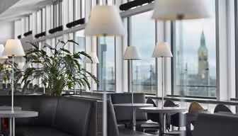 Moderne lounge med sorte lædersæder, sølvborde og hvide lamper. Store vinduer giver udsigt til et sløret klokketårn. Grønne planter bidrager til den afslappede atmosfære.