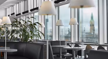 Moderne lounge med sorte lædersæder, sølvborde og hvide lamper. Store vinduer giver udsigt til et sløret klokketårn. Grønne planter bidrager til den afslappede atmosfære.